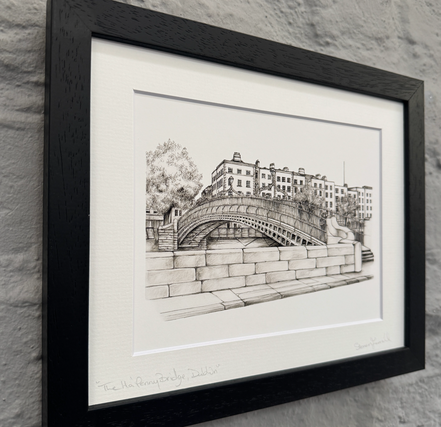 Framed black and white drawing of a bridge and buildings on a stone wall.