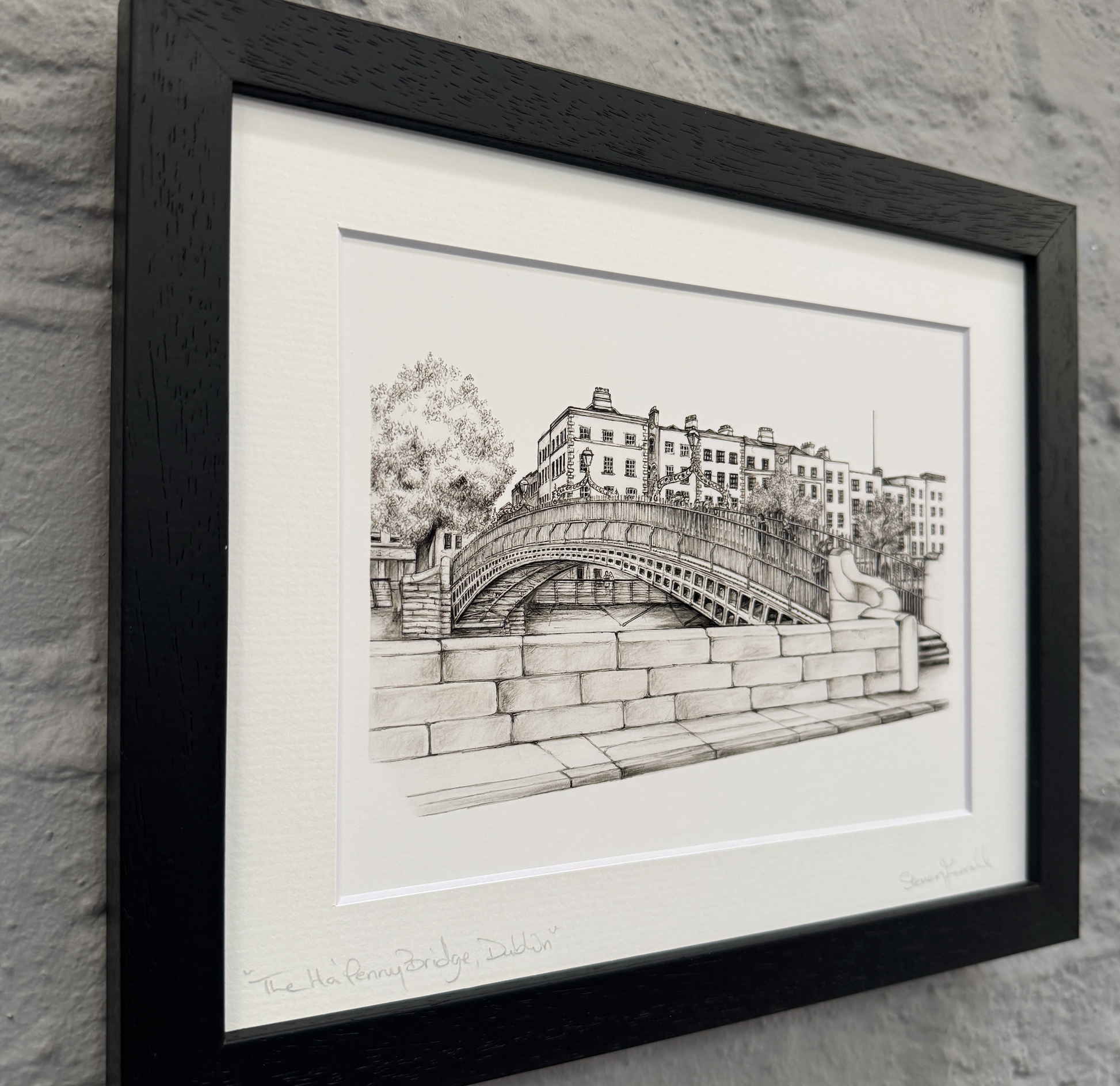 Framed black and white drawing of a bridge and buildings on a stone wall.